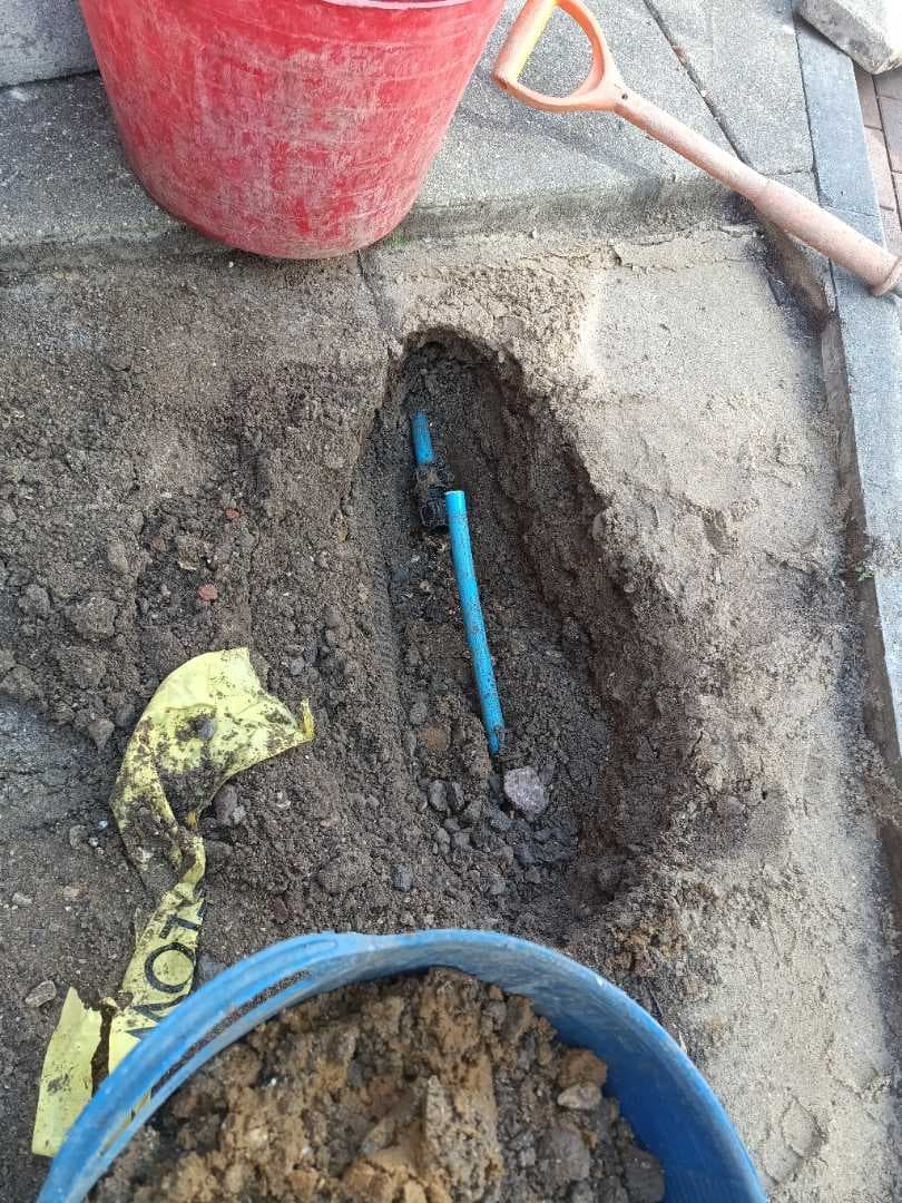A blue plastic water pipe exposed in a narrow trench dug through paving, with buckets and a spade alongside