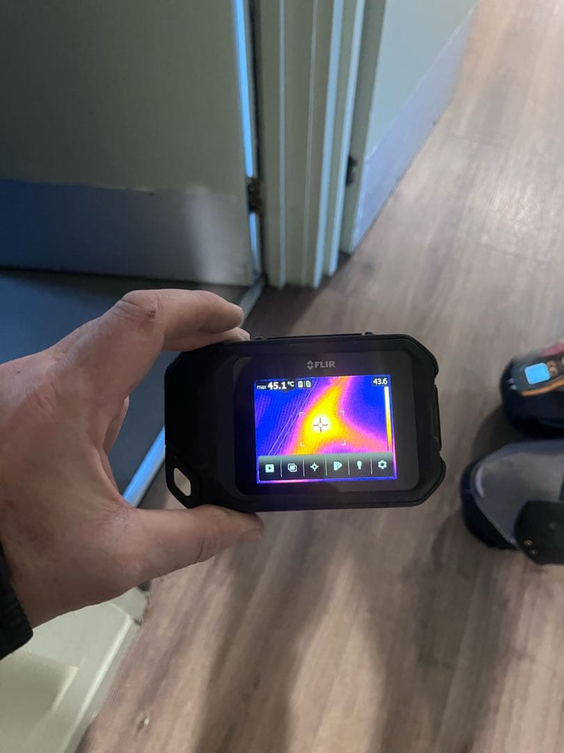 A hand holding a thermal camera over a laminate floor, its screen showing a bright hotspot